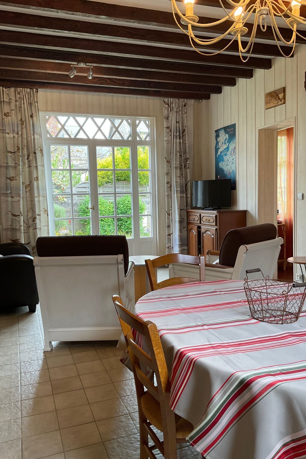 A dining area with a table set for two, a television on a stand, and a view of greenery outside.