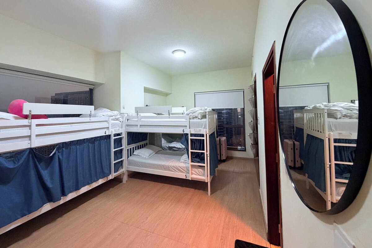 A dormitory room with bunk beds, a large mirror, and a window with blinds.