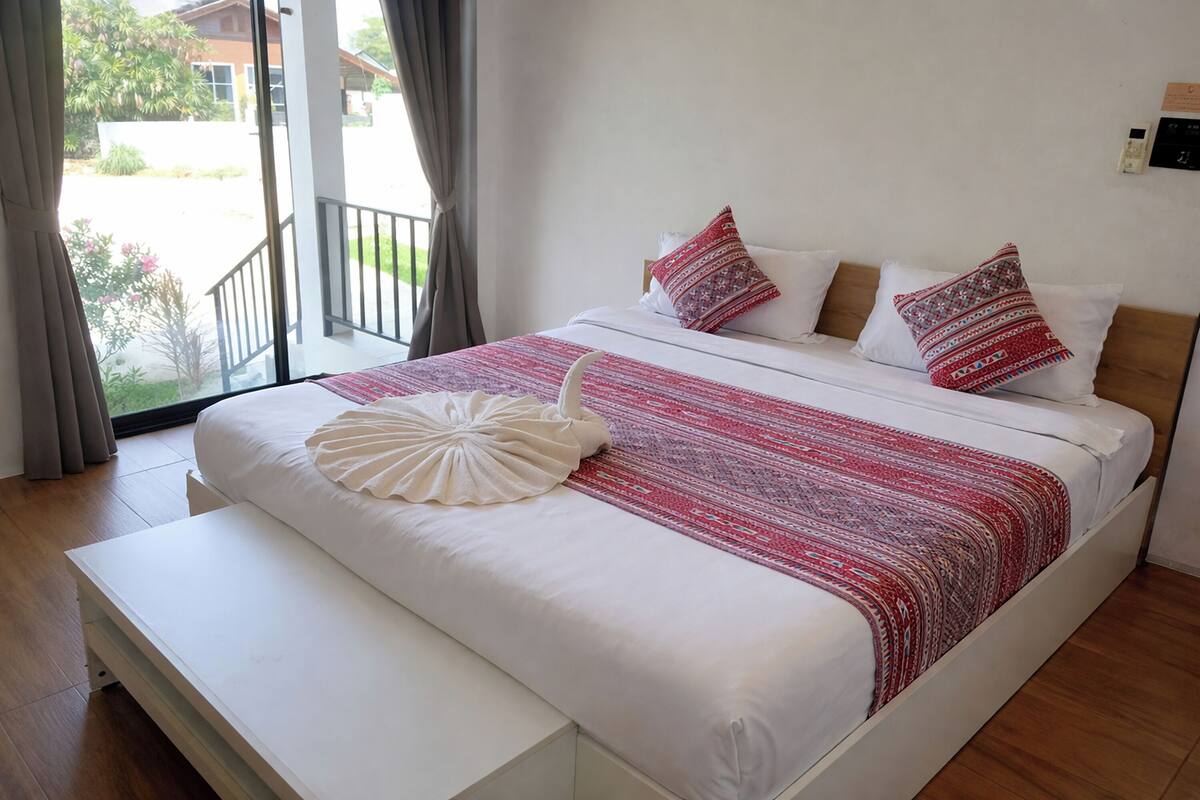 A bedroom with a bed, a bench, a window with curtains, and a view of the outside.