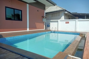 A rectangular swimming pool with clear blue water, surrounded by a wooden deck and a white wall with a window.