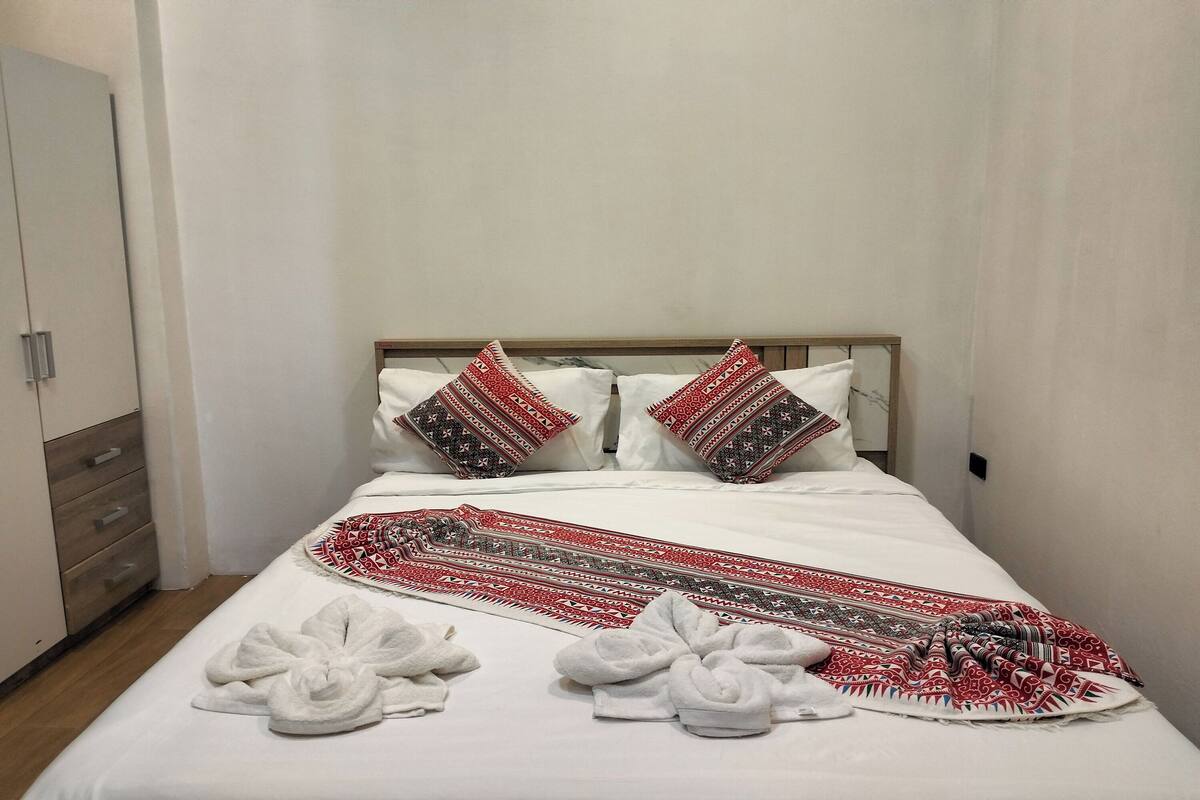 A neatly made bed with a patterned blanket, pillows with a red and black design, and a white towel arrangement on the side.
