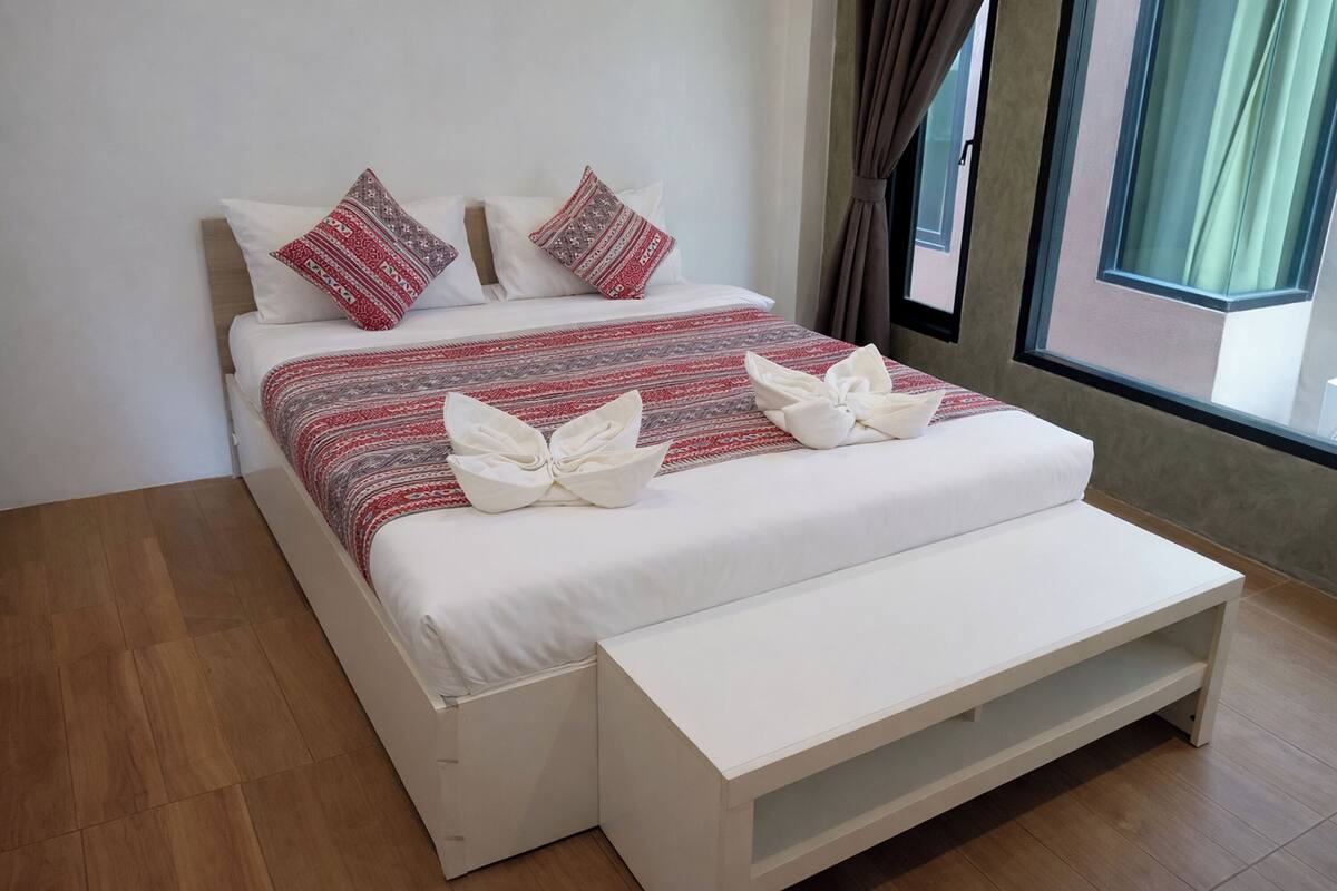 A neatly made bed with patterned pillows, a white bedside table, and a window with curtains.