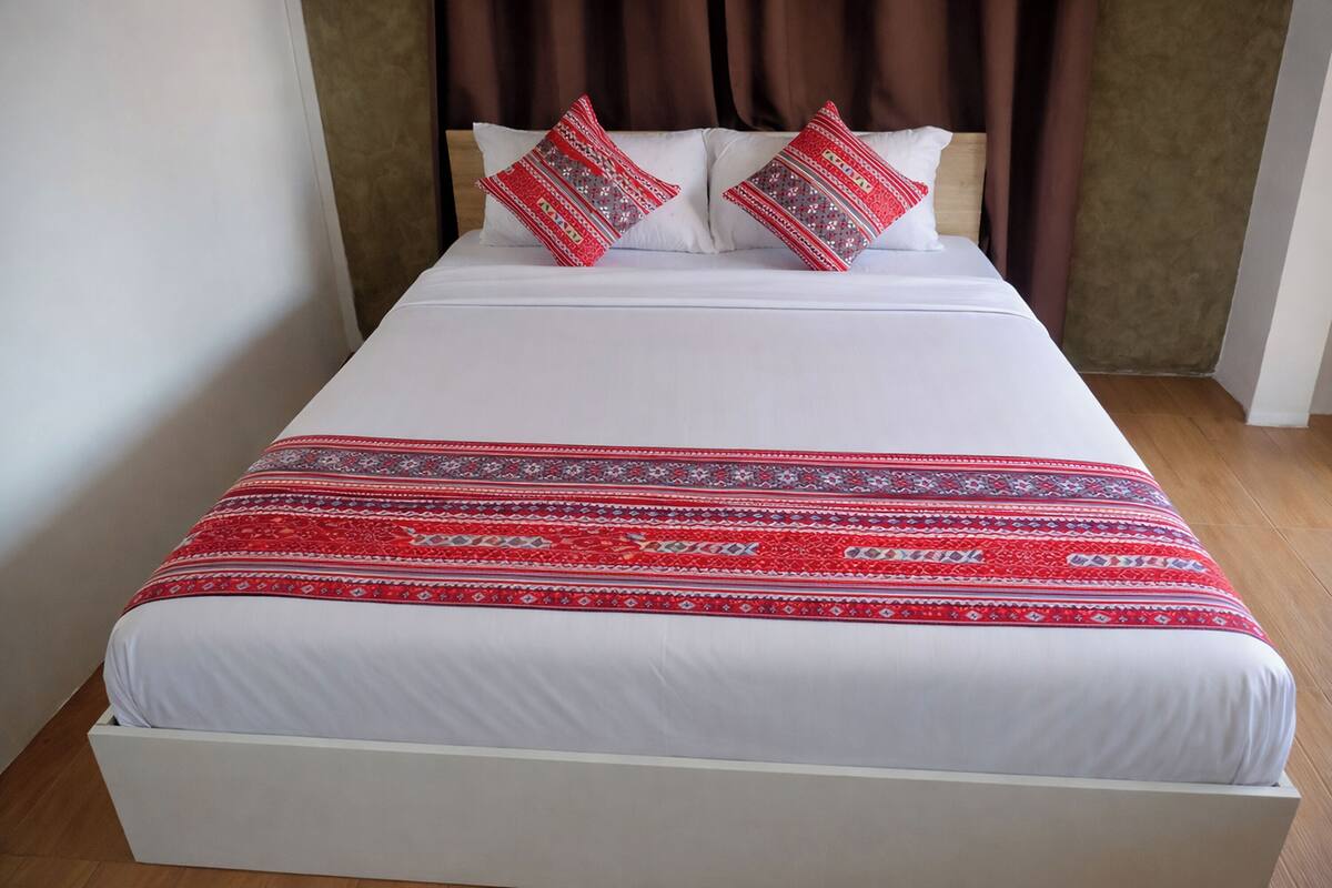 A bed with white bedding and a red patterned blanket, two decorative pillows, and a wooden headboard.