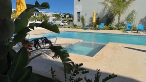 Un área de piscina con sillones de descanso y sombrillas, rodeada de vegetación y un edificio moderno.