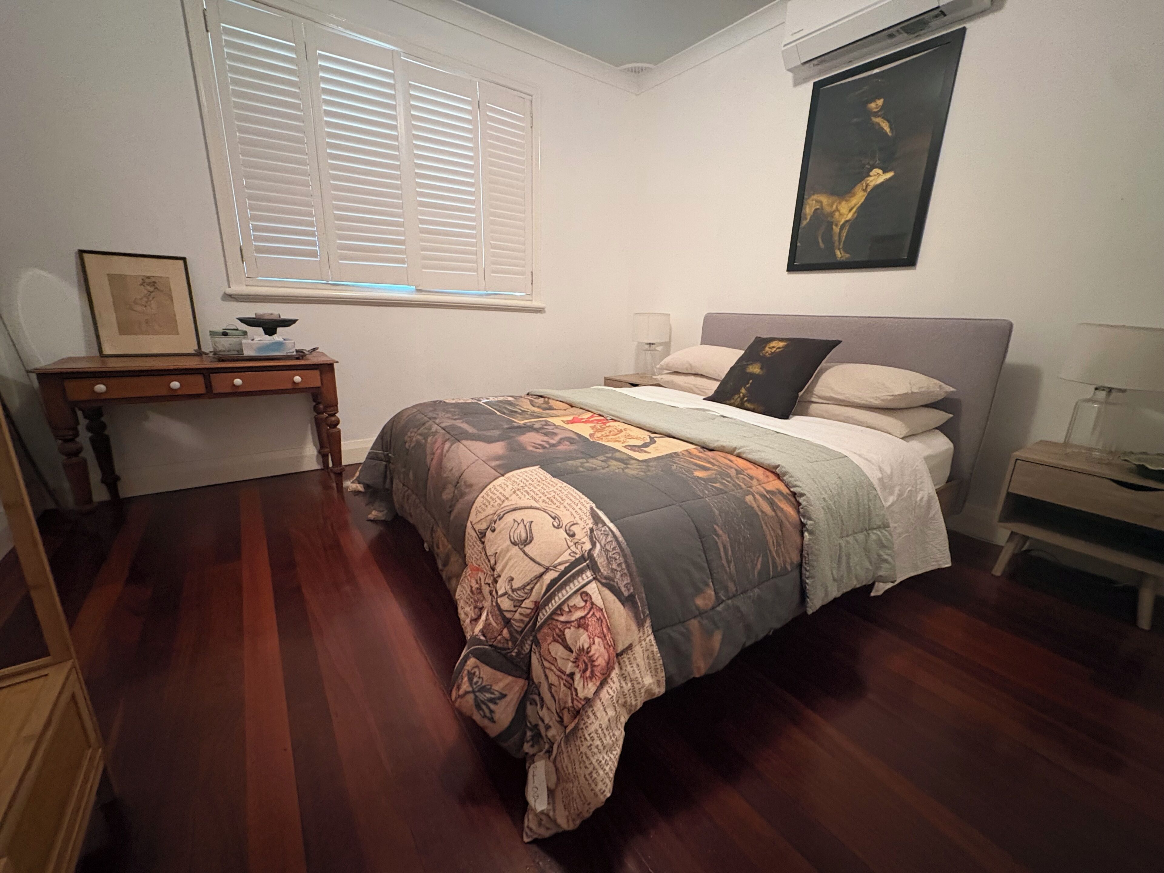A bedroom with a bed, a desk, a window with blinds, a nightstand, and a wall-mounted picture.