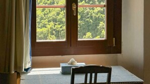 Quarto com porta de madeira, janela com vista para um morro arborizado, mesa com caixa de lenços e uma cadeira.