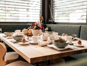 A dining table set with various bowls, cups, and a basket of bread, placed in a room with a large window and a green sofa. - Apart  Bieles NEU by Interhome (Kauns)