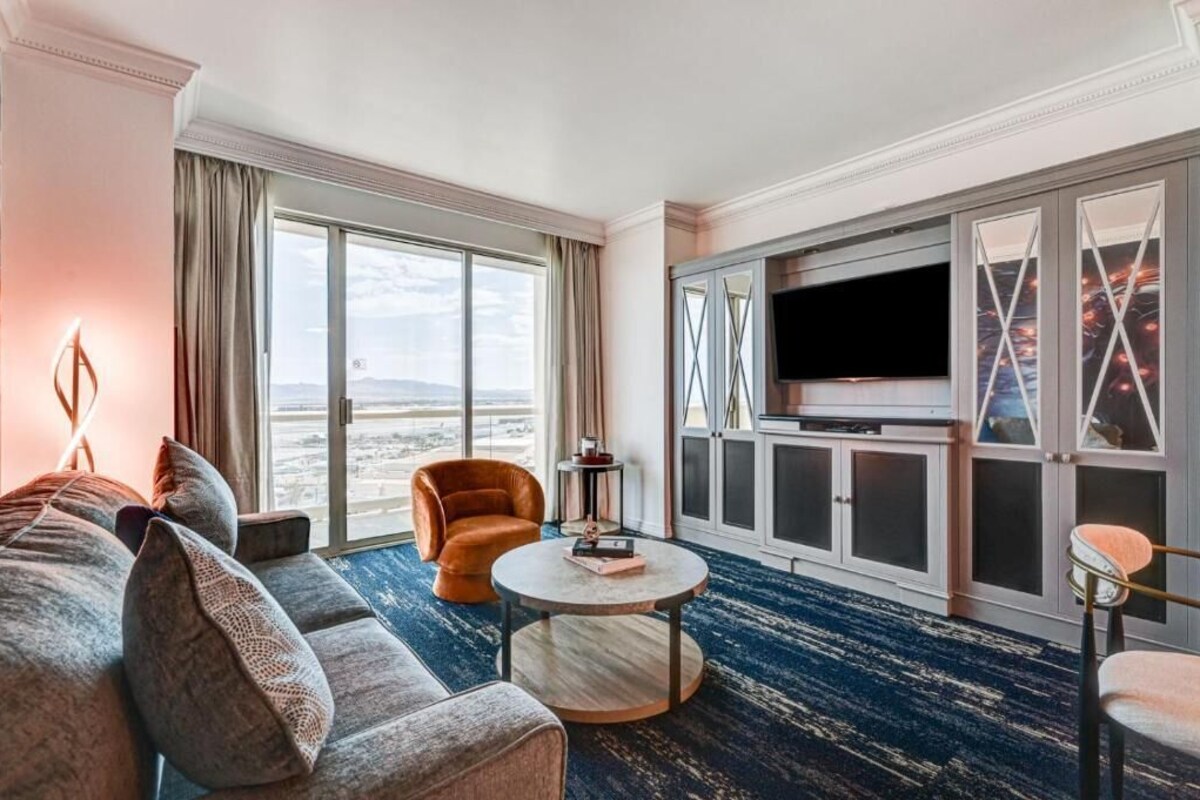 A hotel room with a large window, a sofa, a round coffee table, and a television on a cabinet.