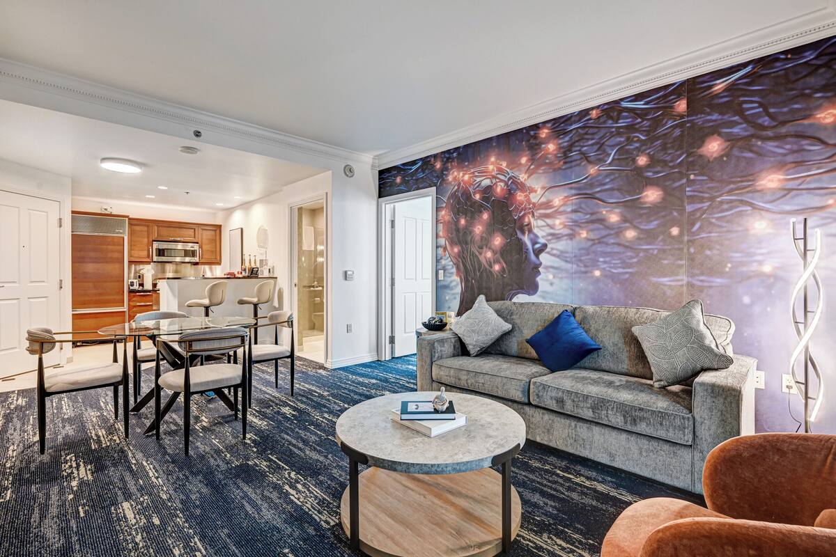 A modern hotel room with a dining area, a gray sofa with multiple pillows, a round coffee table, and a large wall mural.