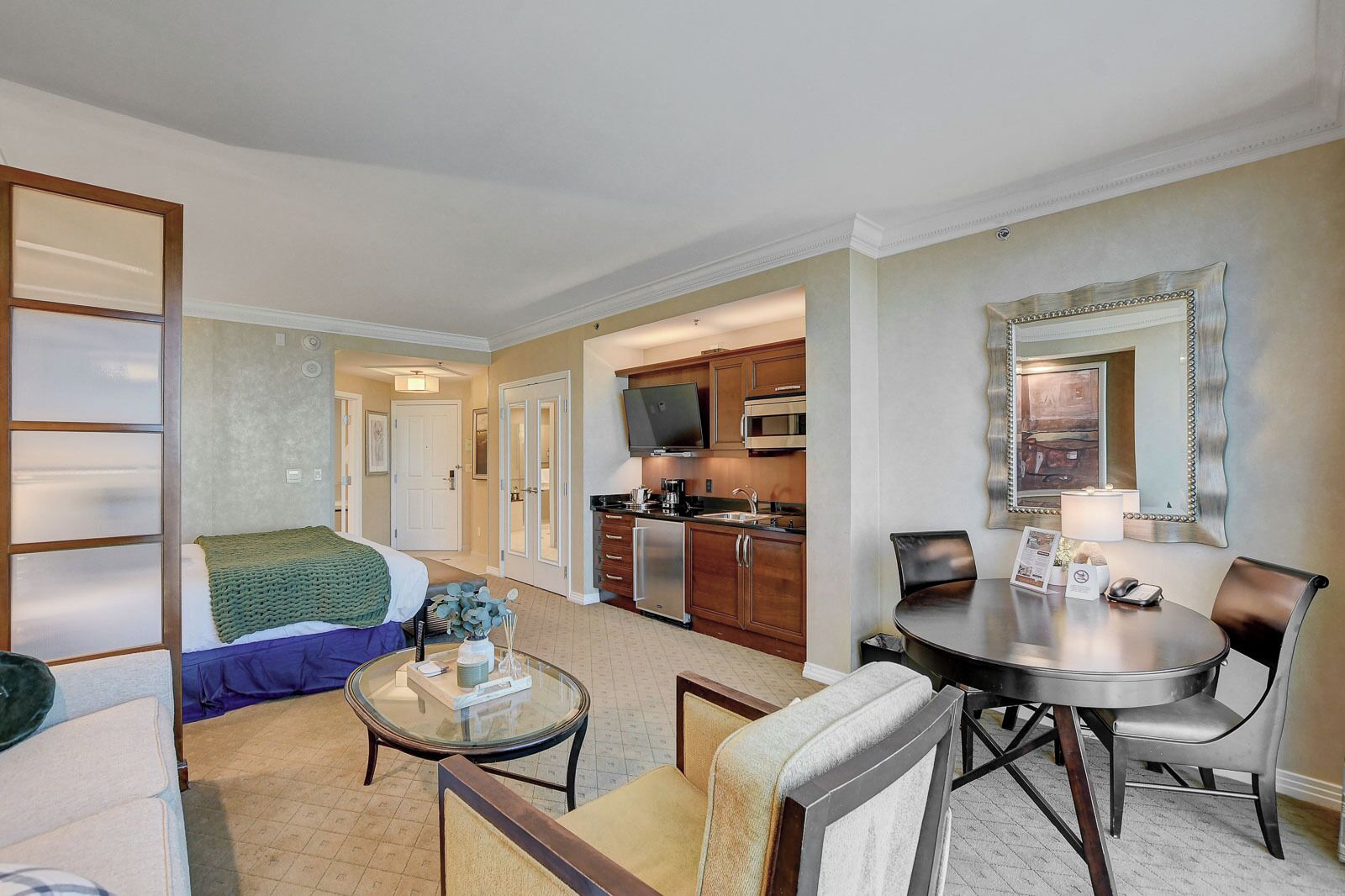 Habitación de hotel con cama, una mesita de vidrio, una mesa de comedor con sillas, zona de cocina con armarios y electrodomésticos, y un espejo grande en la pared.