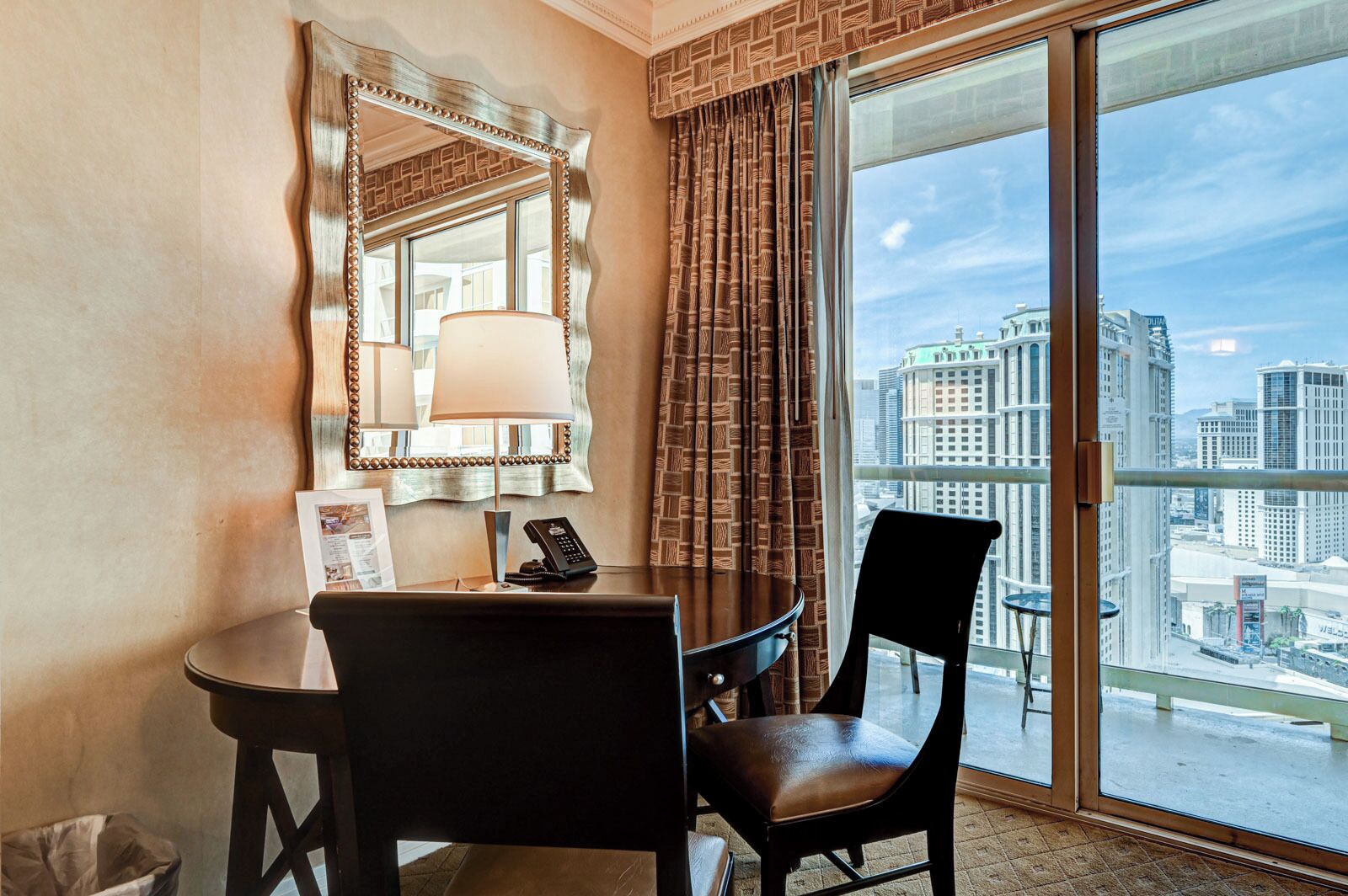 A hotel room with a large window, a round table with a chair, a telephone, and a mirror.