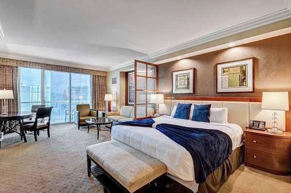 A hotel room with a large bed, a desk, two chairs, a small table, and a view of the city. - Deluxe Balcony Strip View MGM Signature (Las Vegas)