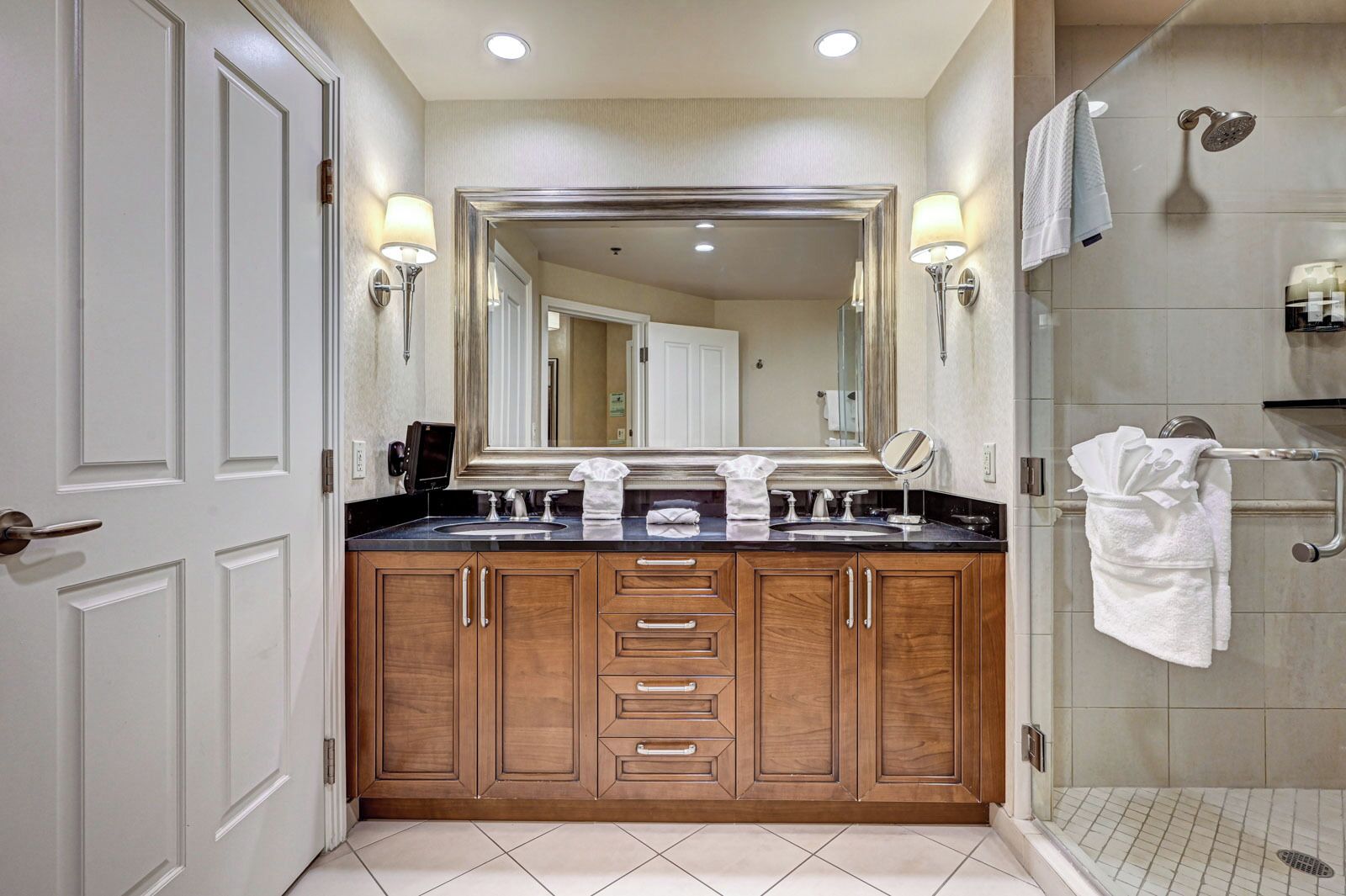 Un baño moderno con un espejo grande, muebles de madera, encimera negra, zona de ducha y un perchero para toallas.