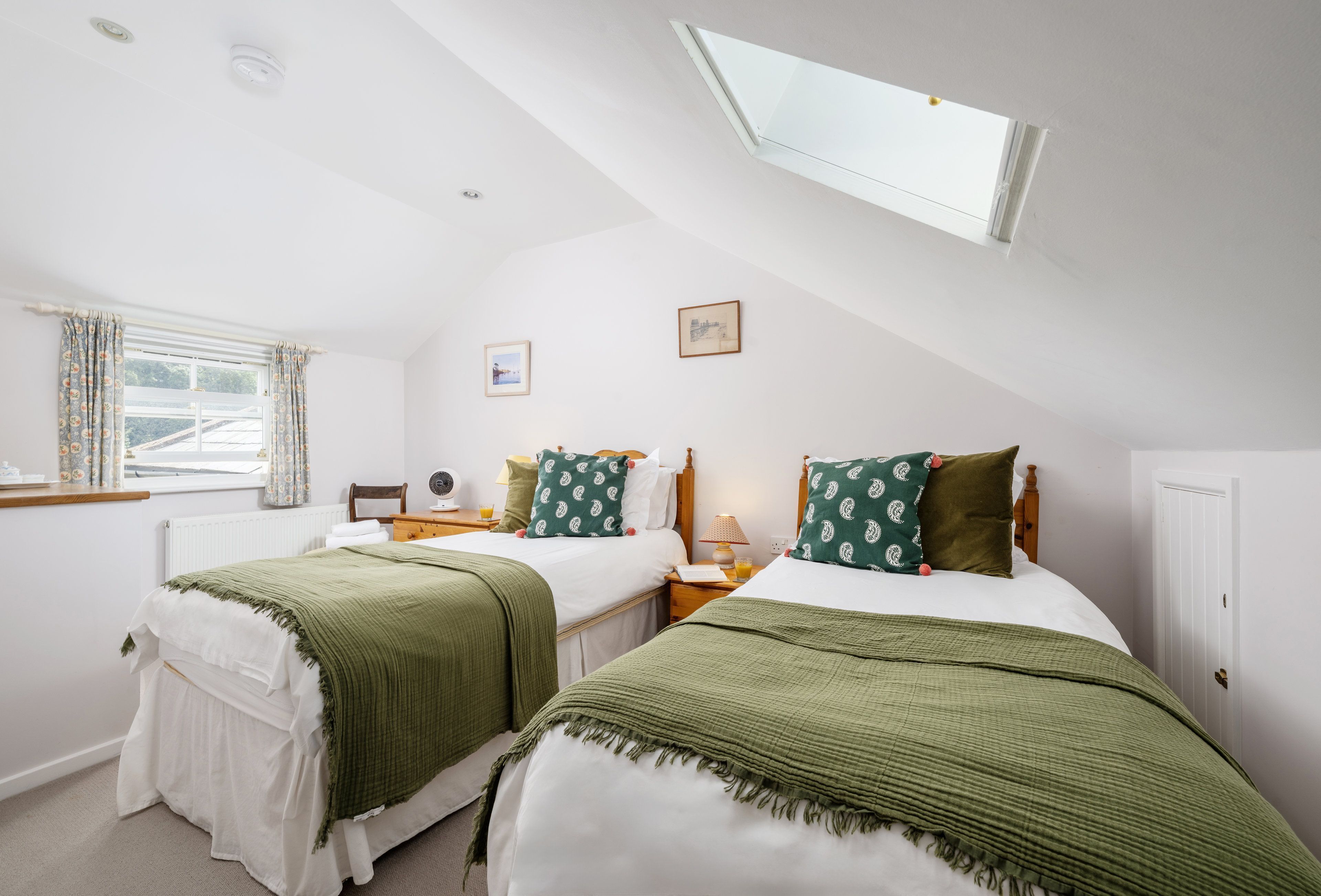 A bedroom with two beds, a window with curtains, a bedside table with a lamp, and a picture on the wall.