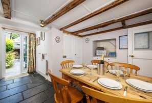Un coin repas avec une table en bois dressée pour quatre personnes, une cuisine équipée d’un four à micro-ondes et une vue sur le jardin par la porte ouverte.