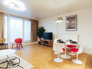 Ein modernes Wohnzimmer mit Essbereich, einem Flachbildfernseher, einem großen Fenster mit Vorhängen und einem Stadtbild als Wandbild.