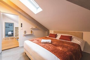 A bedroom with a skylight, a bed with red and white bedding, a wooden bench, and a small shelf with items. - MM Suites City House Cathedrale (Reims)