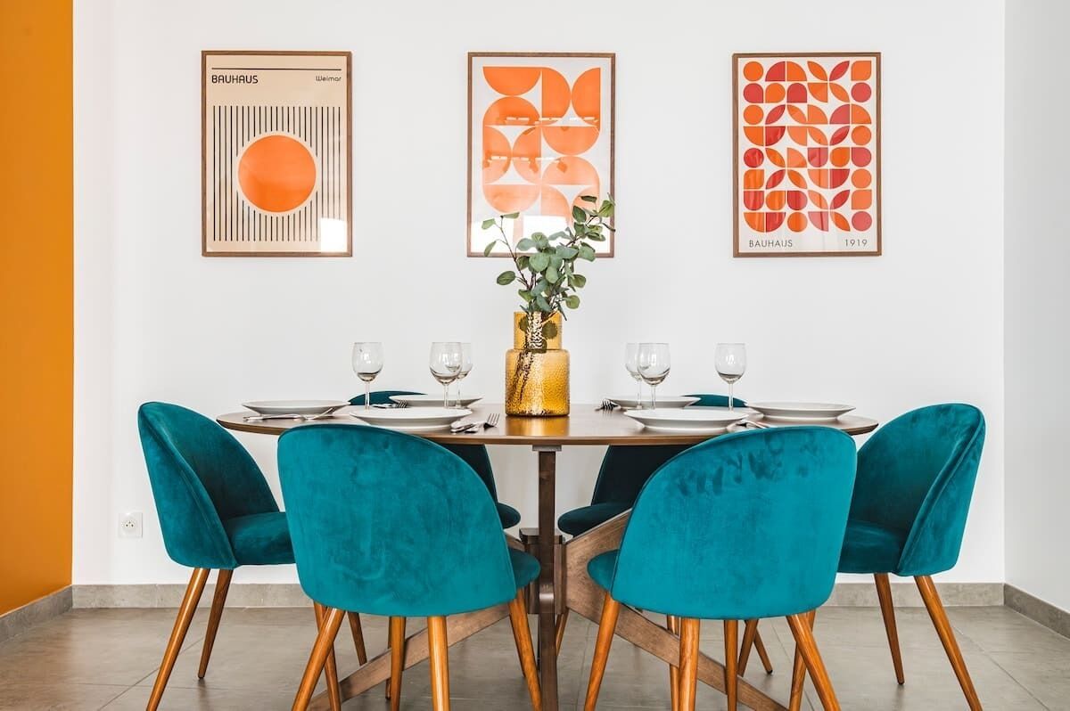 Un comedor con una mesa preparada para cuatro personas, sillas color turquesa y cuadros en la pared.