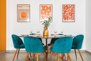 A dining area with a table set for four, teal chairs, and framed artwork on the wall. - MM Suites City House Cathedrale (Reims)