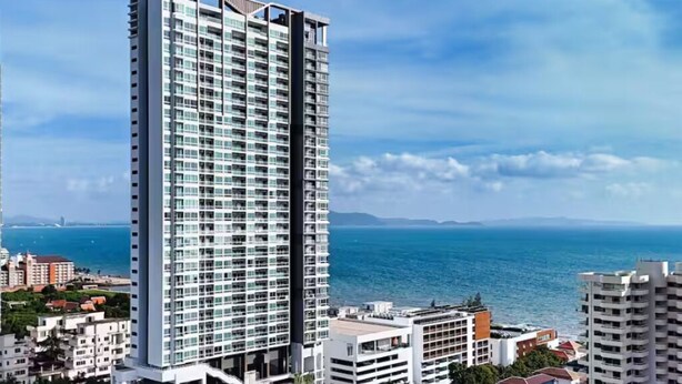 A tall, modern high-rise building with a glass facade, surrounded by other buildings and a body of water in the background. - two-bedroom Beachfront at Veranda Residence (Sattahip)