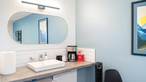 Une salle de bain moderne avec un lavabo blanc, un grand miroir, une machine à café et une poubelle.