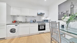 Ett modernt kök med vita skåp, en tvättmaskin, en ugn och en matplats med ett glasbord och stolar.