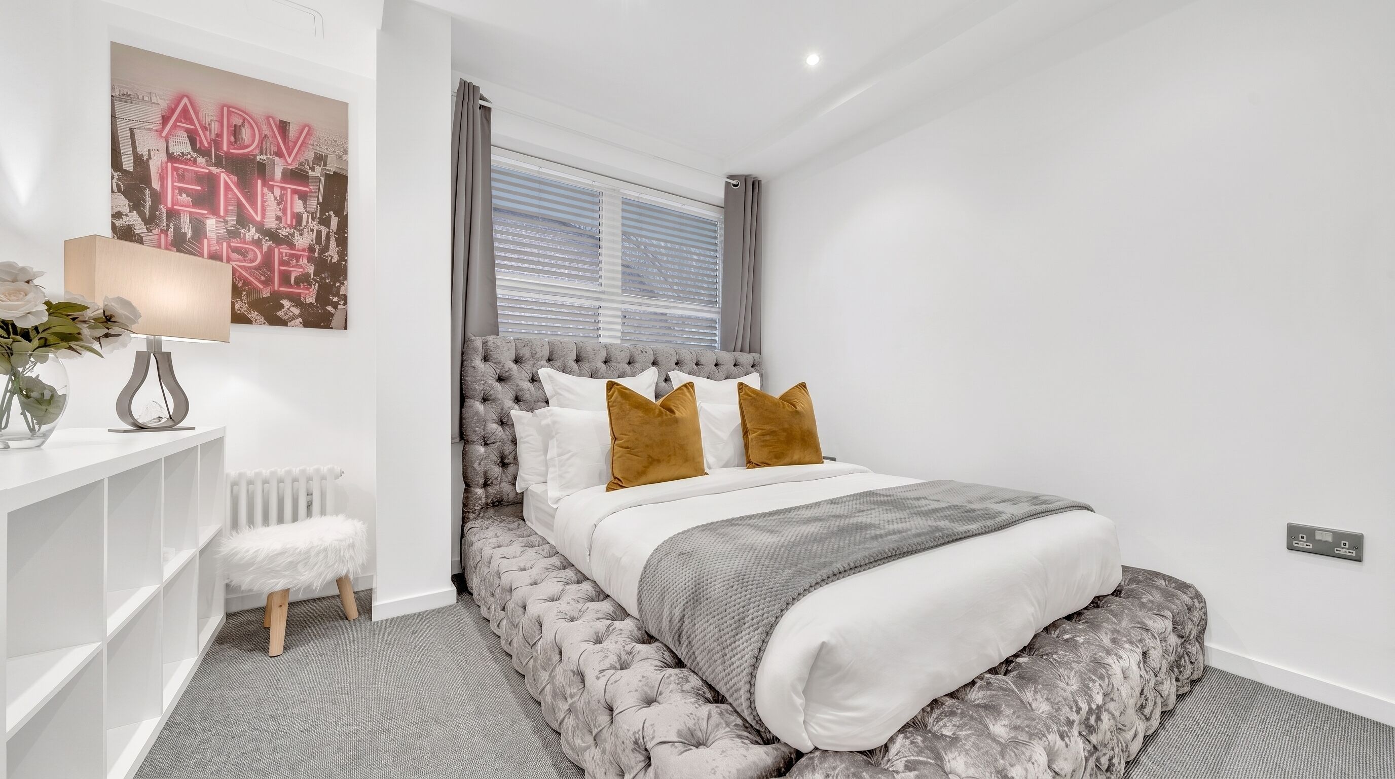 A modern bedroom with a tufted headboard, a white bed with brown pillows, a grey throw, and a radiator.