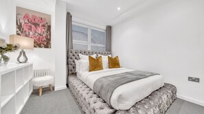A modern bedroom with a tufted headboard, a white bed with brown pillows, a grey throw, and a radiator. - Modern London 1 Bedroom Apartment Camberwell, Oval & Elephant & Castle (LONDON)