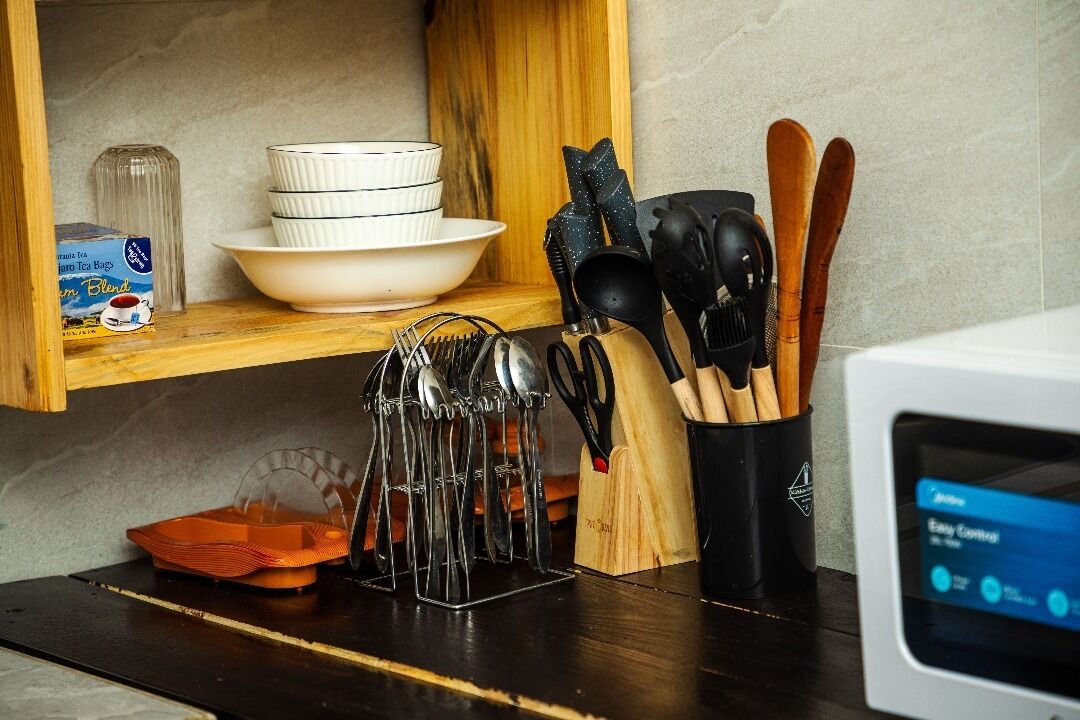A kitchen counter with a microwave, a stack of white bowls, a box of tea bags, a tea kettle, and various utensils.