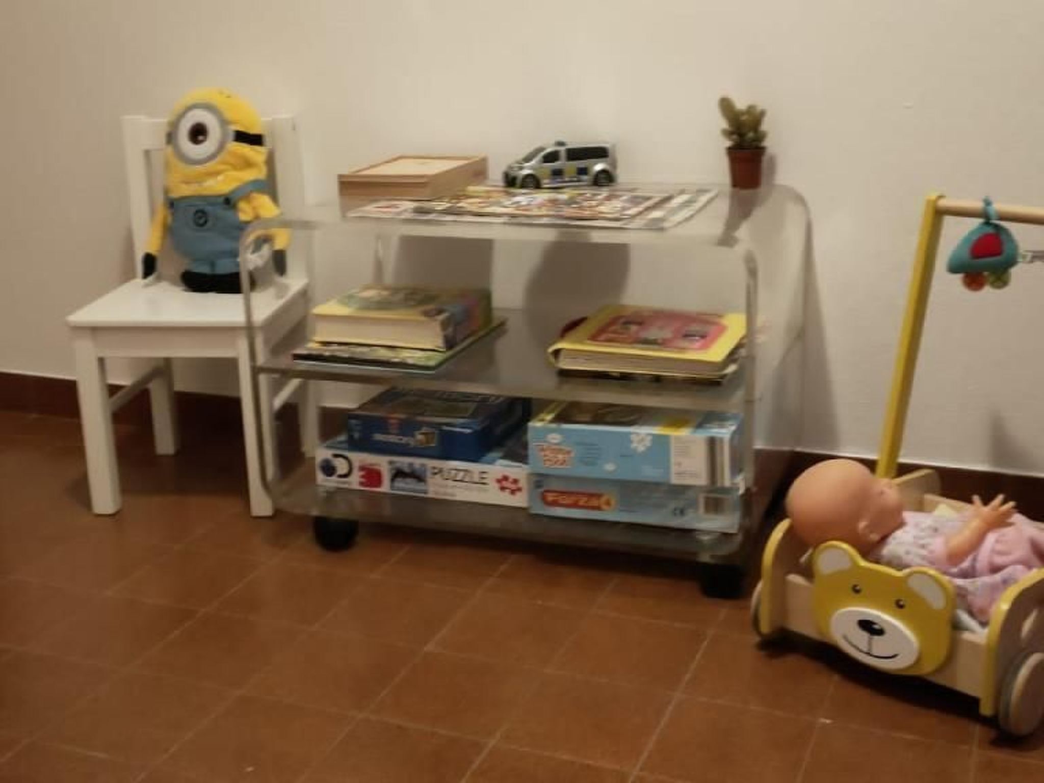 Une pièce avec une chaise blanche, une petite étagère avec des livres et des jouets, et un berceau pour bébé avec un ours en peluche.