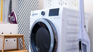 A compact laundry area with a front-loading washing machine, a small wooden stool, and a vacuum cleaner. - "Industrial Sanctum" by Interhome (Brussels)