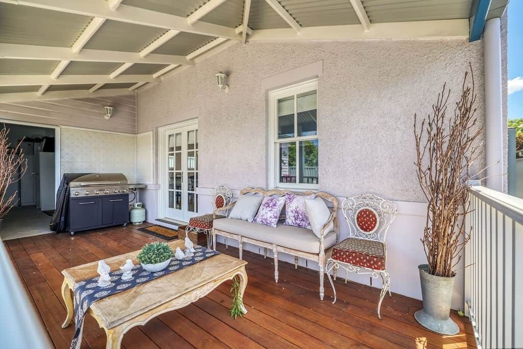 A covered porch with a wooden table, a grill, a bench with cushions, and a potted plant.