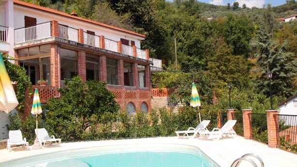Ein Poolbereich mit Liegestühlen und Sonnenschirmen, umgeben von einem Gebäude mit Balkon und Blick auf einen bewaldeten Hügel.
