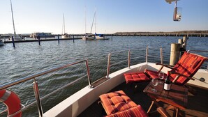 Ein Bootsdeck mit Liegestühlen, einem Tisch, einer Flasche und Gläsern, und Blick auf den Yachthafen mit Segelbooten.