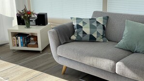 Ein graues Sofa mit gemusterten Kissen, ein weißer Beistelltisch mit einem Radio und Büchern sowie eine Vase mit Blumen.