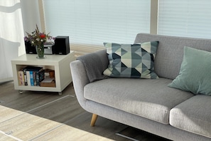 Ein graues Sofa mit gemusterten Kissen, ein weißer Beistelltisch mit einem Radio und Büchern sowie eine Vase mit Blumen.