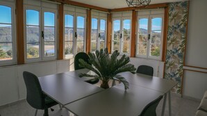 A dining area with a table and chairs, large windows with a view of the outdoors, and a plant centerpiece.