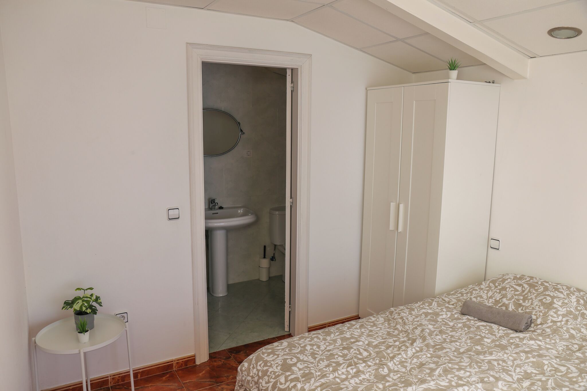Una camera da letto con un letto, un armadio, un tavolino con una pianta e un bagno visibile attraverso una porta aperta.