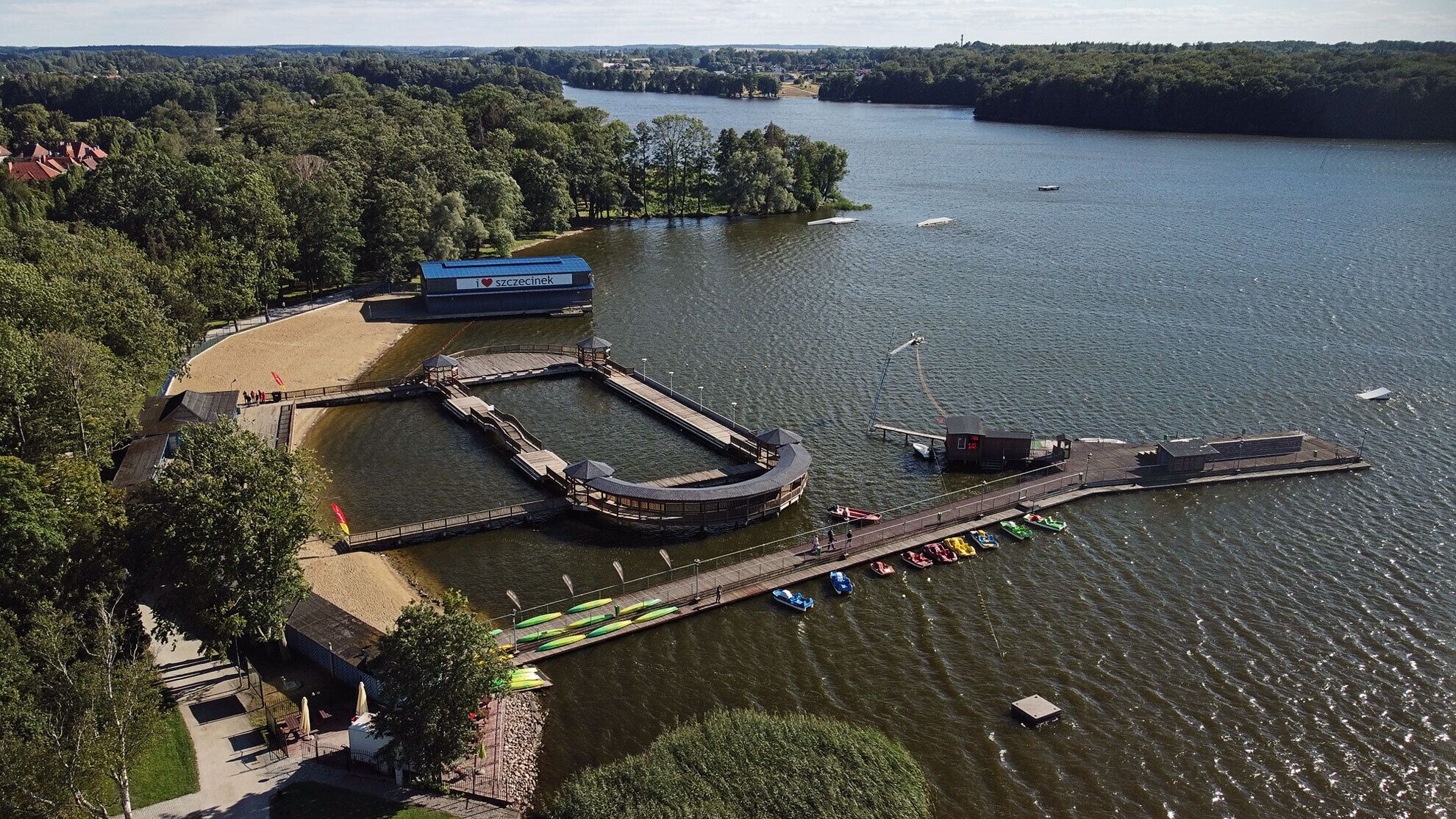 An aerial view of a lakeside park with a dock, boats, and a building labeled 'i love szczecinek'.