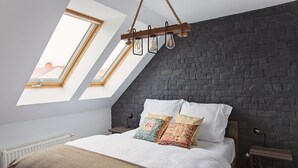A bedroom with a sloped ceiling, skylights, a bed with white linens, and a dark brick wall.
