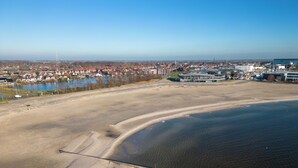 Ein Luftbild einer Küstenlandschaft mit Sandstrand, einem Gewässer und einer in der Ferne liegenden Stadt mit Gebäuden und einem Kirchturm.