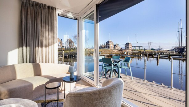 Ein modernes Wohnzimmer mit großem Fenster, einer Couch, einem kleinen Tisch und Blick auf eine Marina mit Booten und Gebäuden.