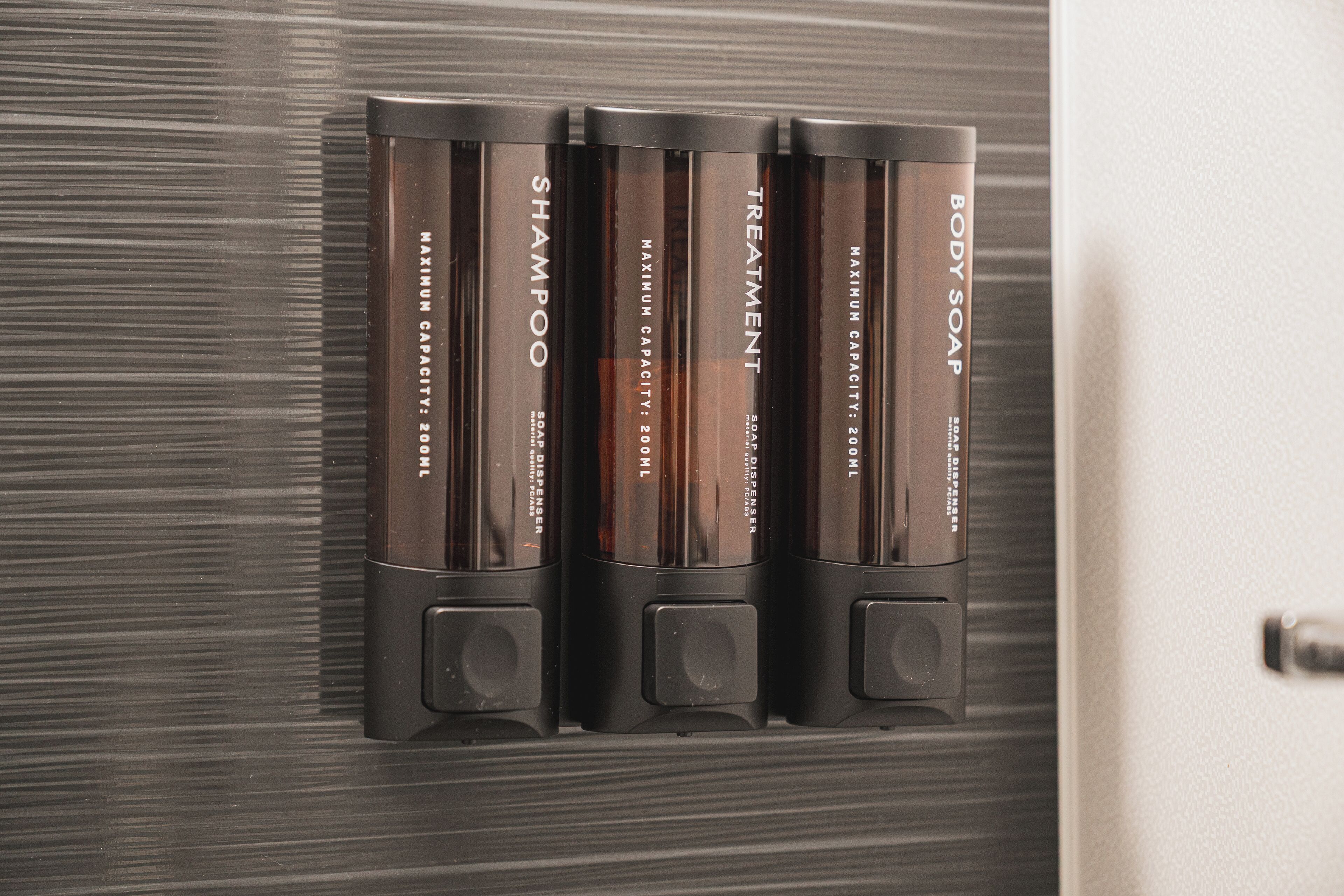 Dispensador de pared con cuatro botellas etiquetadas como 'Champú', 'Tratamiento' y 'Jabón de cuerpo'.
