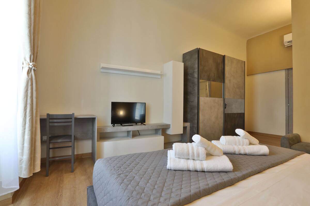A modern hotel room with a bed, a desk, a television, and a wardrobe.