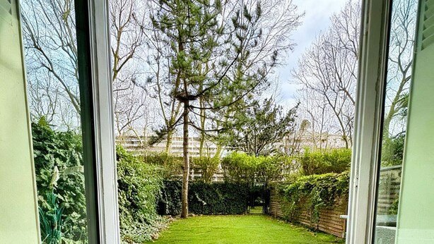 A view through a glass door into a backyard with a lawn, a wooden deck, and a hedge. - Exceptional apartment with private garden (Neuilly-sur-Seine)