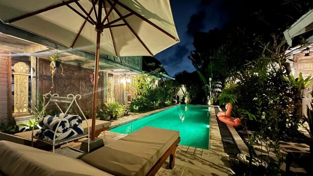 A nighttime pool area with lounge chairs, a swing chair, and an umbrella. - Wooden Cabin in Jimbaran (Jimbaran)
