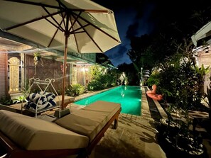 Un espace piscine aménagé pour la nuit, avec des chaises longues, un fauteuil suspendu et un parasol.
