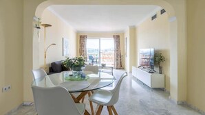 Een moderne eetruimte met een glazen tafel en witte stoelen, een bank en een televisie op een witte kast.
