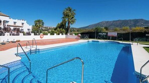 En swimmingpool med kristallklart blått vatten, omgiven av ett vitt poolområde och metallräcken. I bakgrunden syns en vit byggnad med ett rött tegeltak, palmer och ett bergigt landskap.