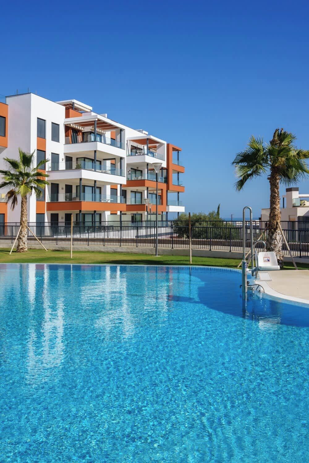 A modern hotel with a swimming pool, multiple balconies, and palm trees.